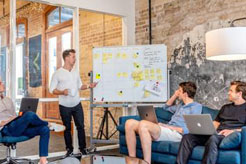 three men sitting while using laptops and watching man beside whiteboard